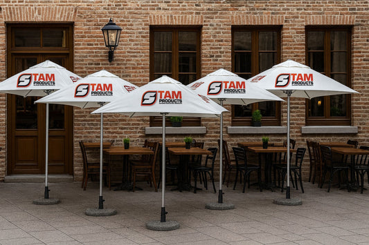 Outdoor setup of five Stam Products custom printed branded white umbrellas with logo, placed over café tables against a brick wall background.